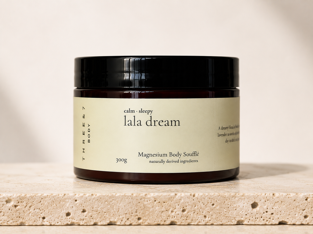 THREE & 7 Body lala dream Magnesium Body Soufflé 300g in a deep brown jar with black lid on beige travertine stone against an off-white studio background.