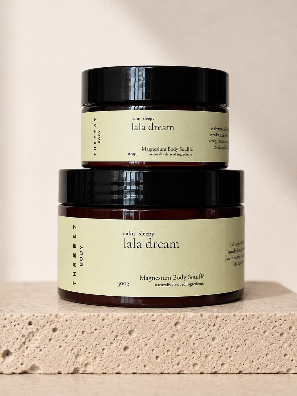 Three & 7 Body lala dream Magnesium Body Soufflé shown in 100g and 300g sizes on beige travertine stone against an off-white studio background.