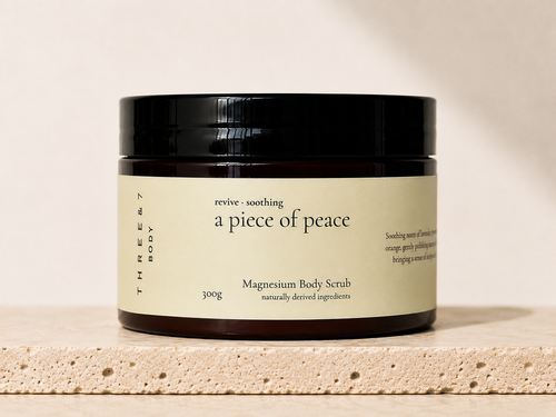 THREE & 7 a piece of peace Magnesium Body Scrub 300g in amber jar on a travertine stone surface.