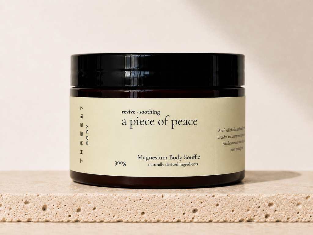 THREE & 7 Body a piece of peace Magnesium Body Soufflé 300g in a deep brown jar with black lid on beige travertine stone against an off-white studio background.