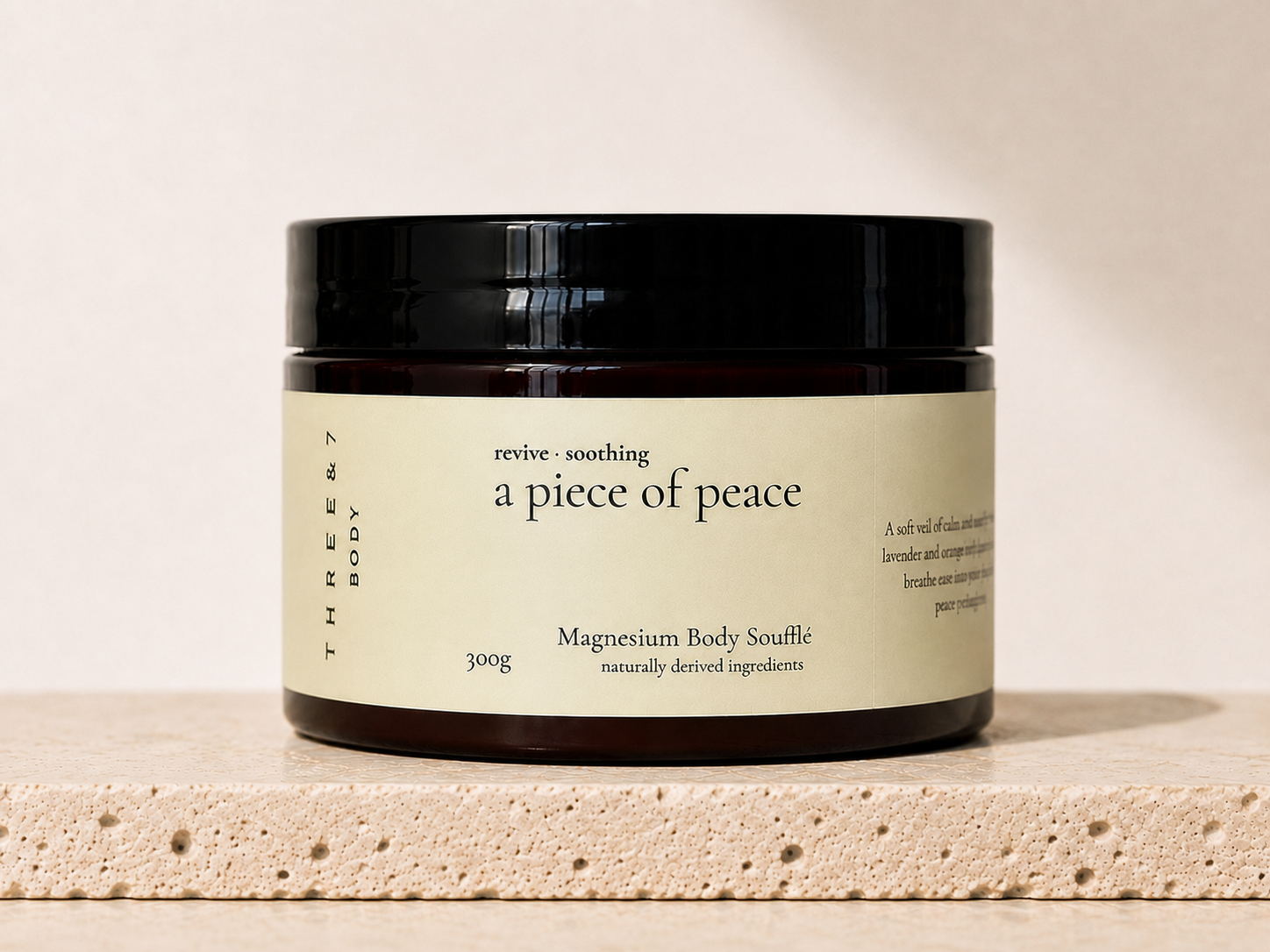 THREE & 7 Body a piece of peace Magnesium Body Soufflé 300g in a deep brown jar with black lid on beige travertine stone against an off-white studio background.