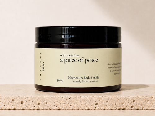 THREE & 7 Body a piece of peace Magnesium Body Soufflé 300g in a deep brown jar with black lid on beige travertine stone against an off-white studio background.