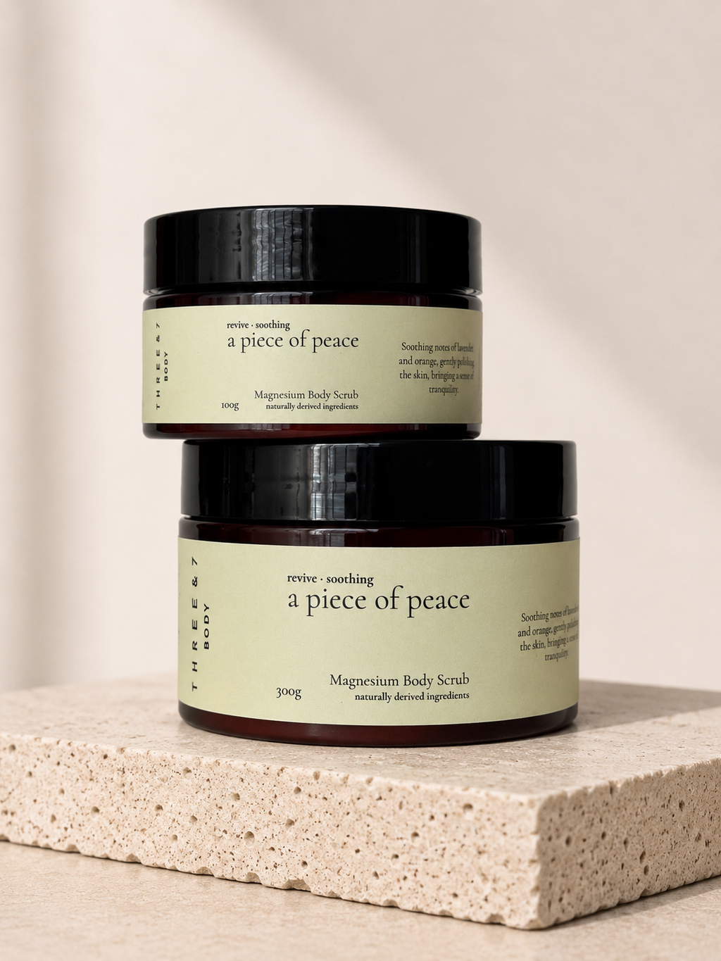 Three & 7 Body a piece of peace Magnesium Body Scrub shown in 100g and 300g sizes on beige travertine stone.