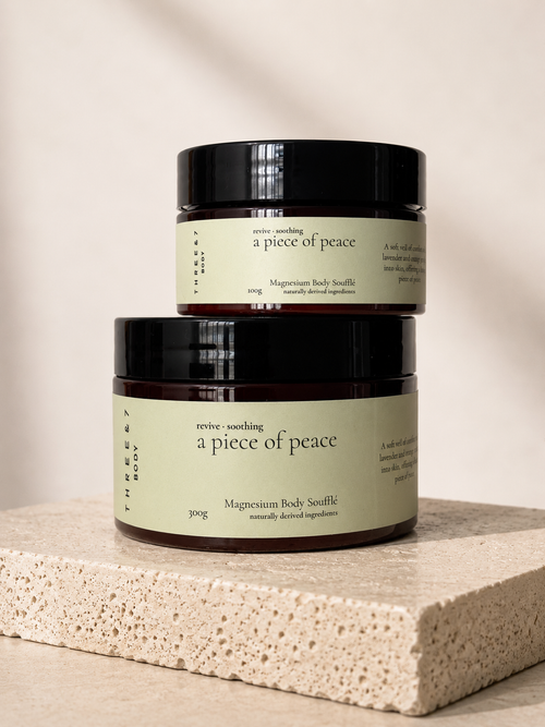 Three & 7 Body a piece of peace  Magnesium Body Soufflé 300g and 100g in a deep brown jar with black lid on beige travertine stone against an off-white studio background.