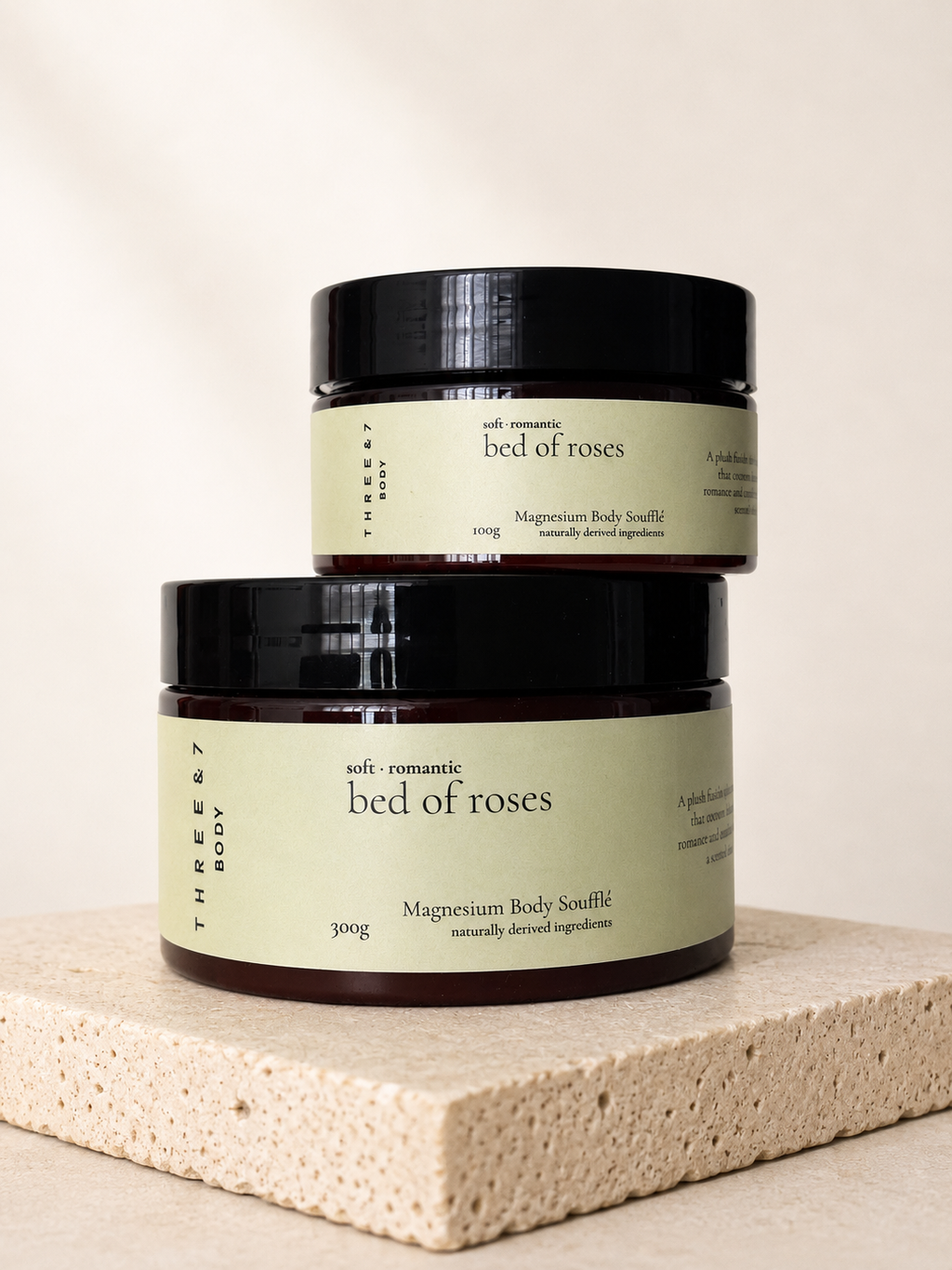 Three & 7 bed of roses Magnesium Body Soufflé shown in 100g and 300g sizes on beige travertine stone.