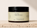 THREE & 7 Body bed of roses Magnesium Body Soufflé 300g in a deep brown jar with black lid on beige travertine stone against an off-white studio background.