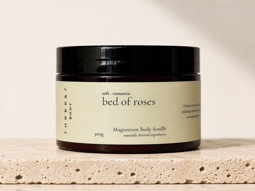 THREE & 7 Body bed of roses Magnesium Body Soufflé 300g in a deep brown jar with black lid on beige travertine stone against an off-white studio background.