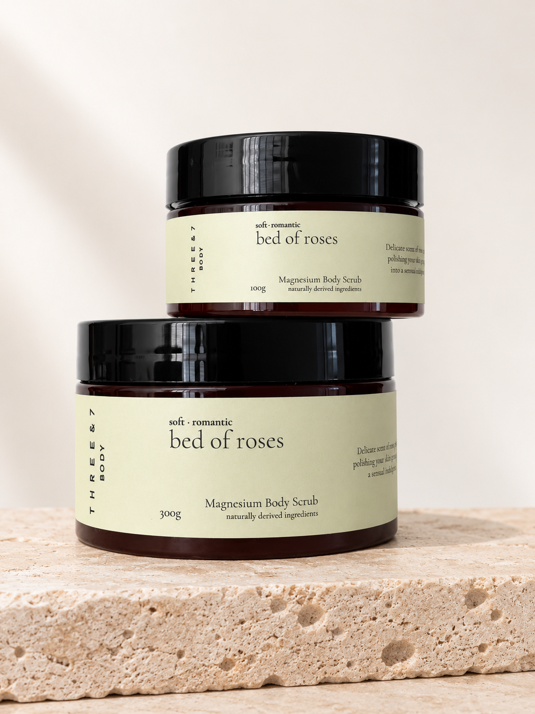Three & 7 Body bed of roses Magnesium Body Scrub shown in 100g and 300g sizes on beige travertine stone.