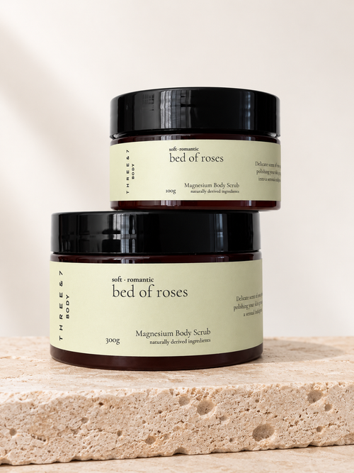 Three & 7 Body bed of roses Magnesium Body Scrub shown in 100g and 300g sizes on beige travertine stone.