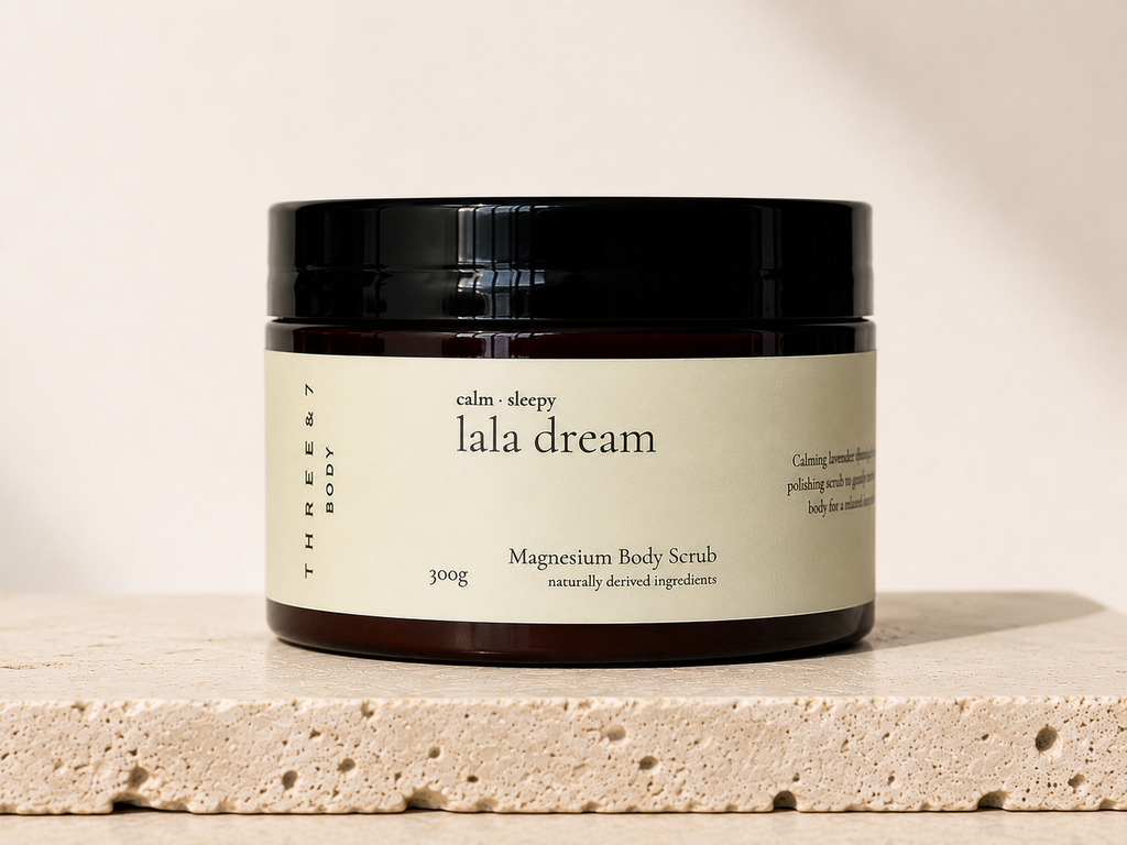 Three & 7 Body lala dream Magnesium Body Scrub 300g on beige travertine stone against an off-white studio background.