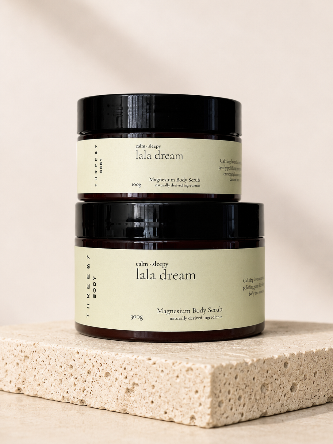 Three & 7 Body lala dream Magnesium Body Scrub shown in 100g and 300g sizes on beige travertine stone.